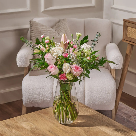 Soft pink and white floral arrangement by Gilded Blooms with roses, lilies, and greenery in a clear glass vase, displayed on a wooden table in a neutral-toned living room setting.