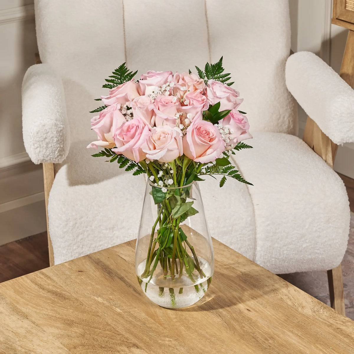 Light pink rose bouquet with baby’s breath and greenery arranged in a clear glass vase on a wooden table in front of a cream boucle chair