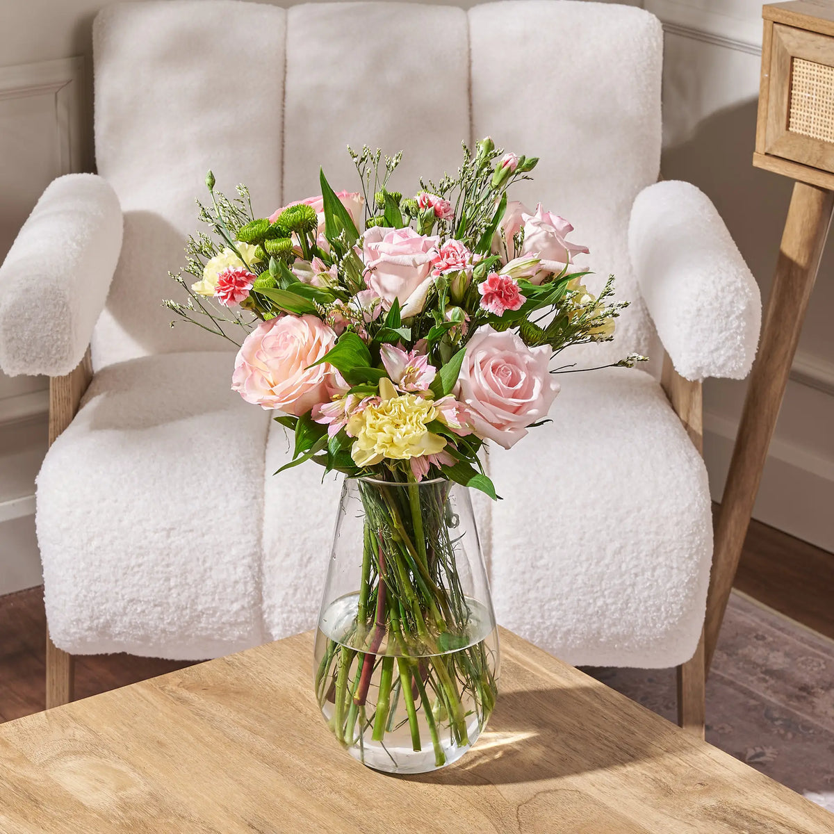 soft pink and ivory rose bouquet with carnations and greenery in glass vase on wooden table with neutral armchair background