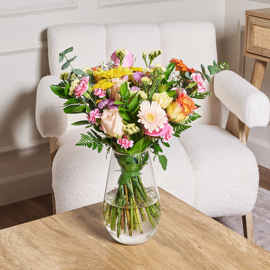 Colourful mixed bouquet featuring pink alstroemeria, pink and red roses, purple daisies, hot pink carnations, pink and purple limonium, ruscus, and leather leaf arranged in a clear glass vase on a wooden table.
