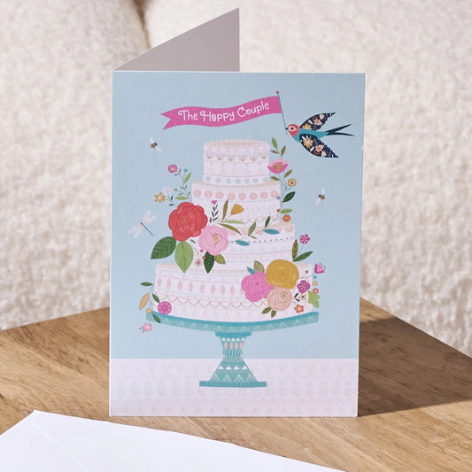 A square greeting card featuring an illustration of a three-tiered decorated cake on a stand, with flowers, bees, a bird, and a pink banner reading 'The Happy Couple'. The card is shown standing on a wooden surface with a textured cream background.