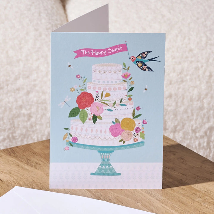 A square greeting card featuring an illustration of a three-tiered decorated cake on a stand, with flowers, bees, a bird, and a pink banner reading 'The Happy Couple'. The card is shown standing on a wooden surface with a textured cream background.