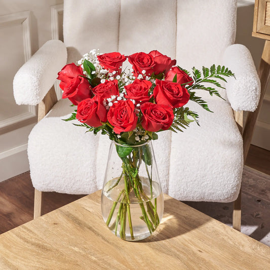 Bouquet of red roses in a clear vase on a wooden table with a white cushioned chair in the background.