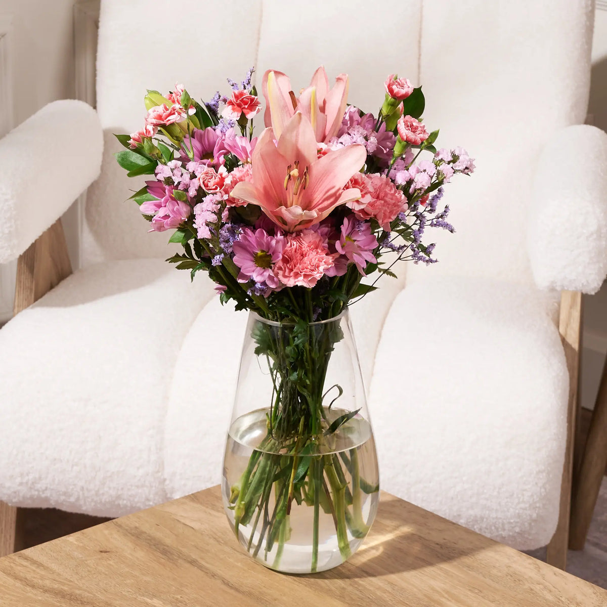 Bouquet of pink and purple flowers by Gilded Blooms in a clear vase on a wooden table with a white chair in the background.