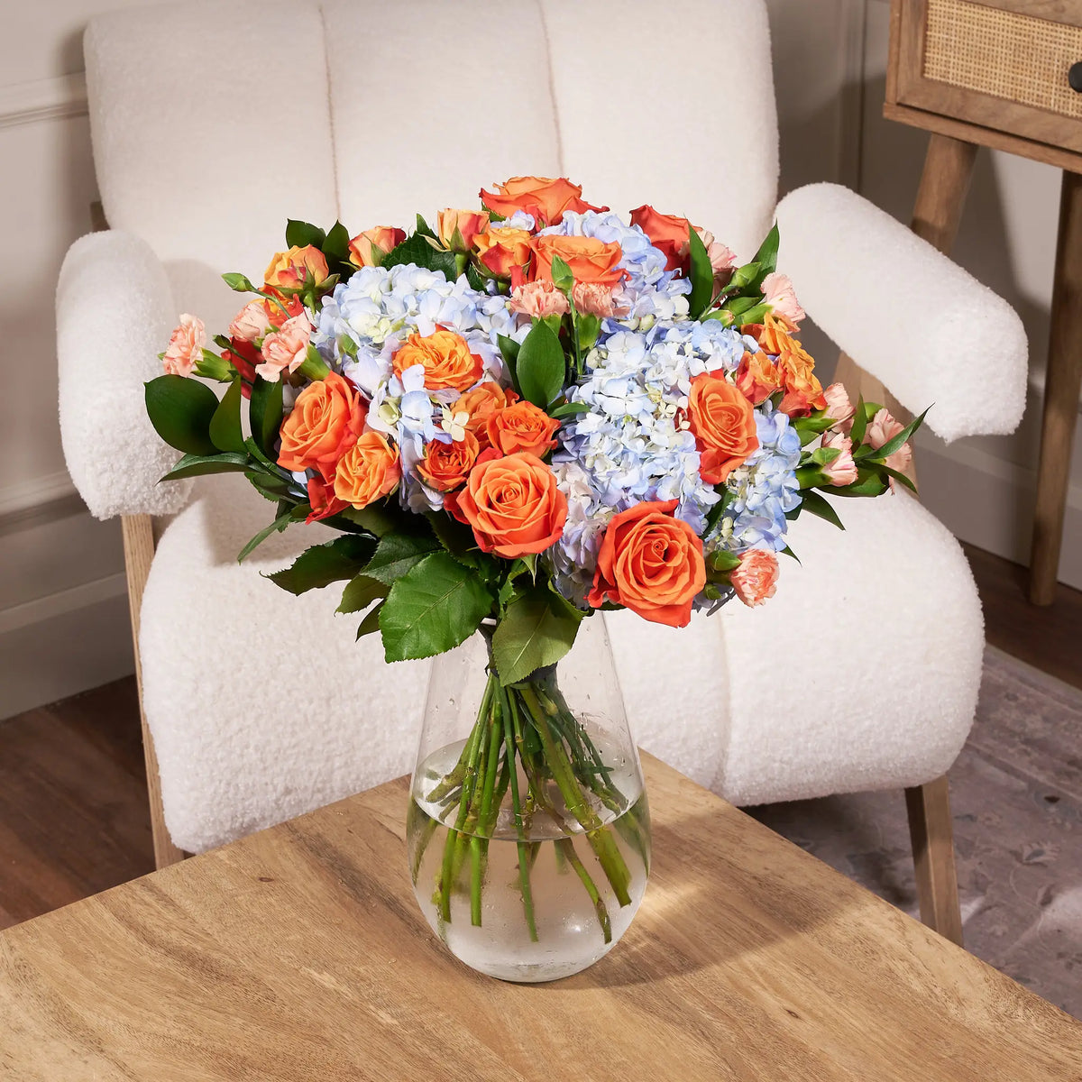 Orange rose and blue hydrangea bouquet featuring peach mini carnations and fresh greenery arranged in a clear glass vase on a wooden table in front of a white accent chair.