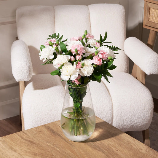 Soft pink and white carnation bouquet featuring mini carnations, ruscus, leather leaf, and fresh greenery arranged in a clear glass vase on a wooden table in front of a white accent chair.