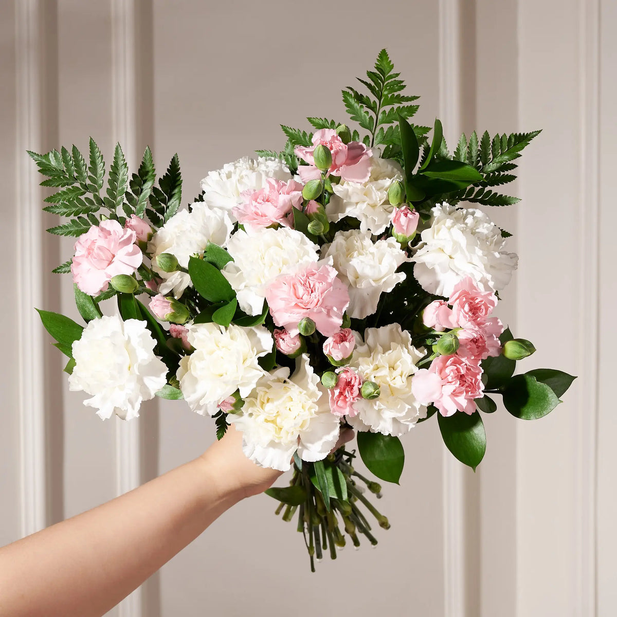 A bouquet of artificial carnations and greenery held by a person's hand against a soft beige paneled background.