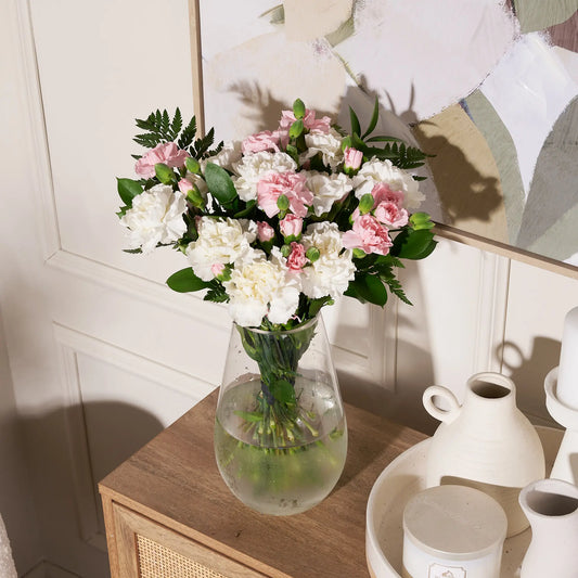 A bouquet of artificial pink and white carnations with green foliage in a clear glass vase, styled on a wooden side table next to white ceramic vases against a cream paneled wall.