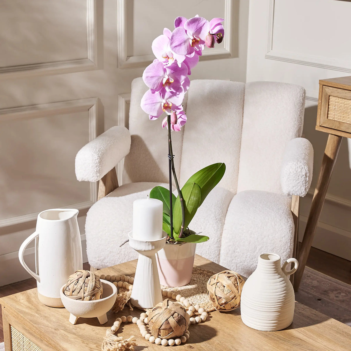 Decorative setting with a white armchair, potted orchid, and various home decor items on a wooden table.