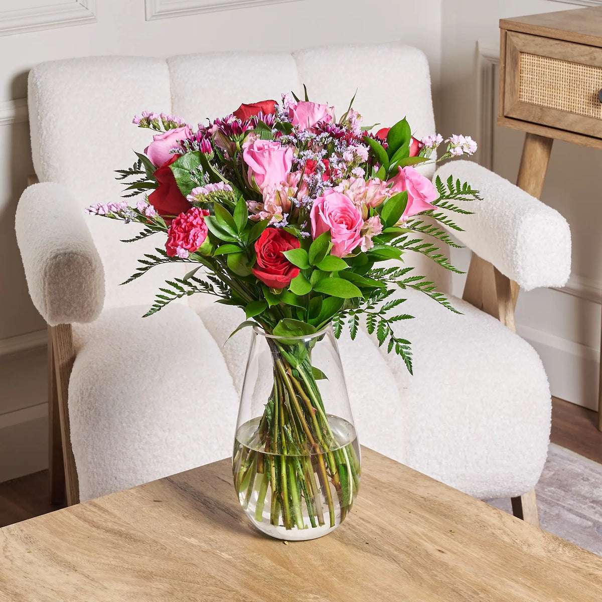 Pink and red rose bouquet featuring roses, alstroemeria, carnations, limonium, ruscus, and leather leaf arranged in a clear glass vase on a wooden table in front of a white accent chair.