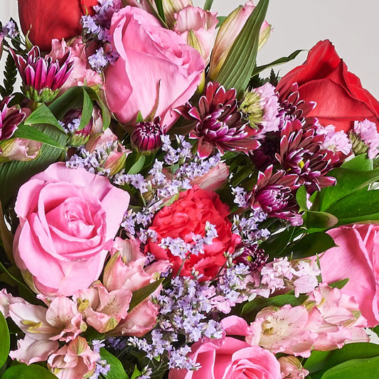 Close-up of The Valentina Bouquet featuring pink and red roses, pink alstroemeria, hot pink carnations, purple daisies, pink and purple limonium, and lush green foliage.