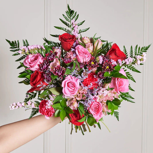 Hand-tied pink and red bouquet featuring roses, alstroemeria, carnations, purple daisies, pink and purple limonium, ruscus, and leather leaf greenery held against a neutral wall.