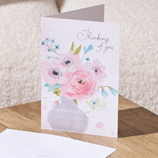 A floral greeting card standing on a wooden surface, with a white envelope in front. The card has a watercolor-style bouquet of pink and blue flowers in a pale lavender vase, with the words 'Thinking of you' printed in script near the top.