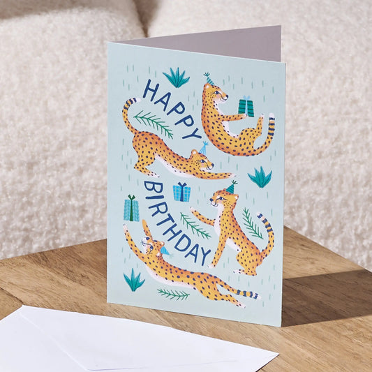 A folded birthday card featuring four playful cheetah illustrations and the words “HAPPY BIRTHDAY,” standing on a wooden surface with a white envelope in front. The background shows a soft beige textured wall.