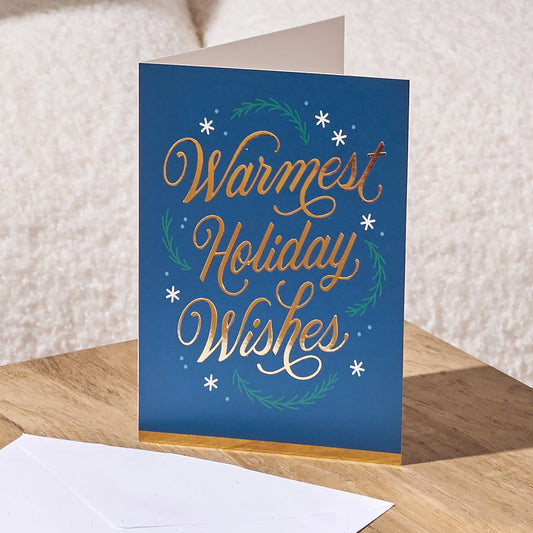A blue greeting card standing on a wooden surface with a white envelope in front. The card reads 'Warmest Holiday Wishes' in metallic gold and white script, decorated with green wreath motifs and small white snowflakes, against a soft beige background.