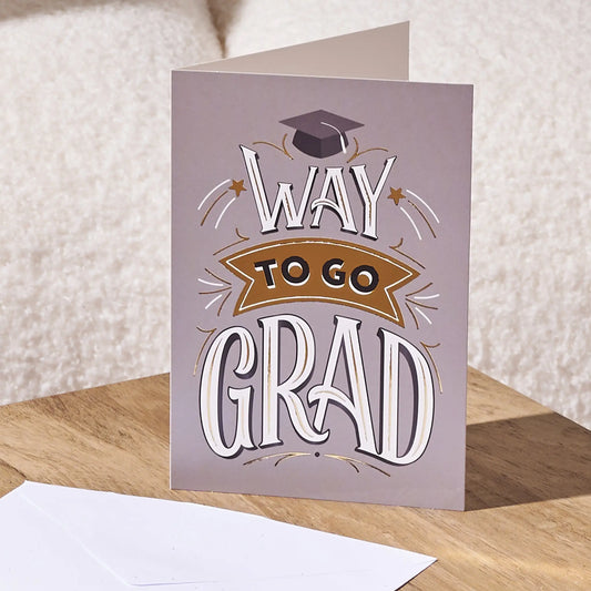 A folded graduation card standing on a wooden surface, with a white envelope in front. The card has a muted lavender-gray background and decorative text reading “WAY TO GO GRAD” with a graduation cap illustration and a tan ribbon banner.
