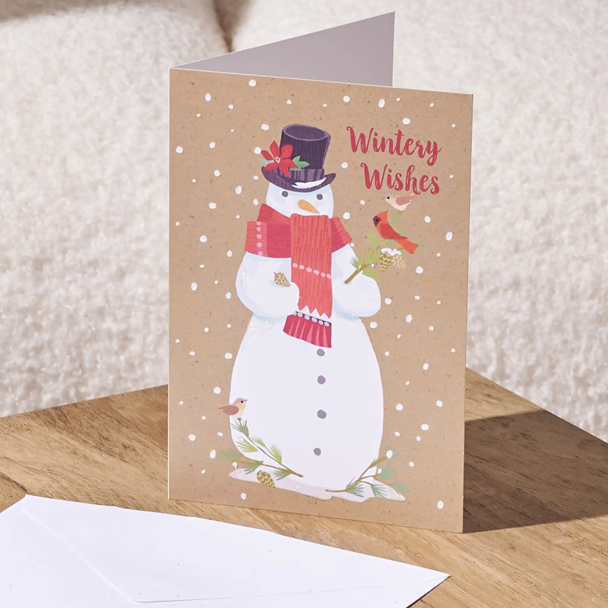 A folded greeting card standing open on a wooden surface, featuring a winter snowman illustration with a red scarf, top hat, birds, and the words 'Wintery Wishes' in red script. A white envelope lies in front, with a soft beige textured background behind.