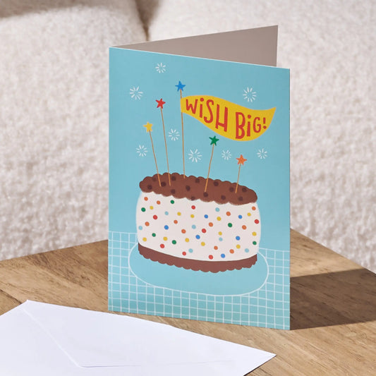 A folded birthday card standing on a wooden surface, showing a light blue card with an illustration of a frosted cake, colorful confetti, star candles, and a yellow banner that reads “WISH BIG!” A white envelope lies in front. The background is a softly blurred beige textured surface.