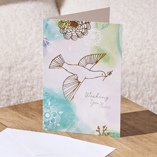 A folded greeting card with a watercolor background in soft teal, mint, and cream, featuring a gold foil dove carrying an olive branch and the words 'Wishing You Peace.' A white envelope lies in front on a wooden surface, with a soft beige textured background behind it.