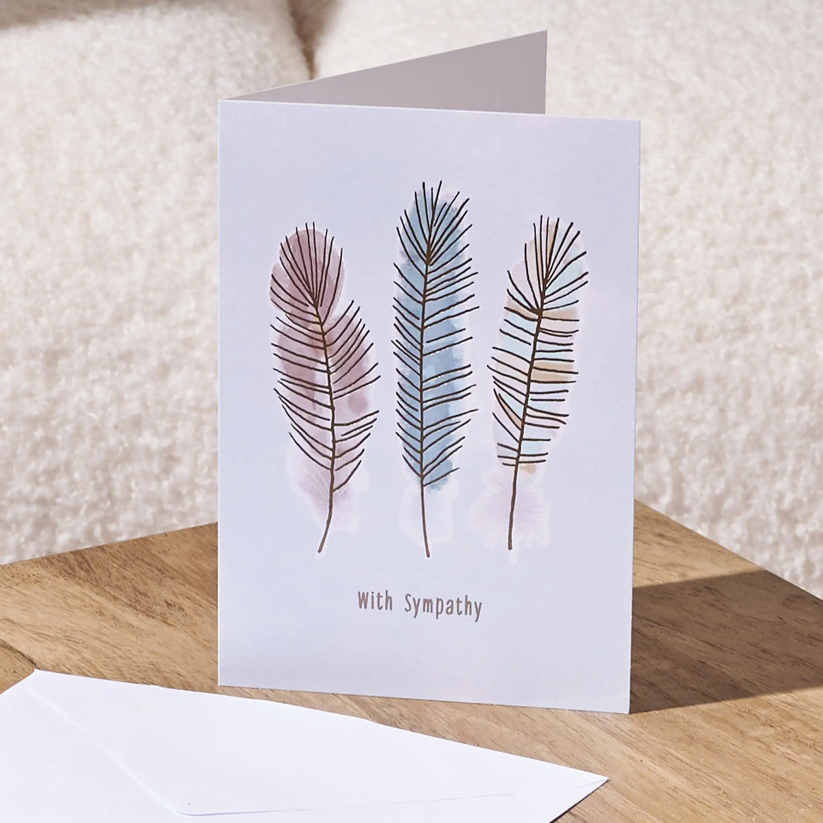 A sympathy greeting card standing on a wooden surface, featuring three illustrated feathers in muted pink, blue, and brown tones above the word 'With Sympathy.' A white envelope lies in front, with a soft cream fabric background behind.