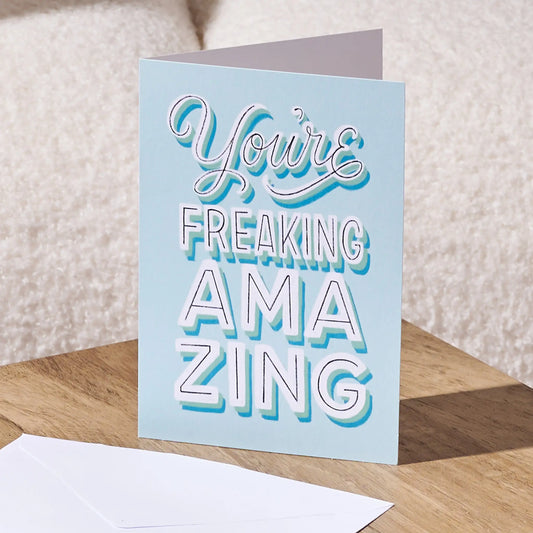 A folded light blue greeting card standing on a wooden surface, with a white envelope in front. The card reads “You’re FREAKING AMAZING” in stylized white and blue lettering.